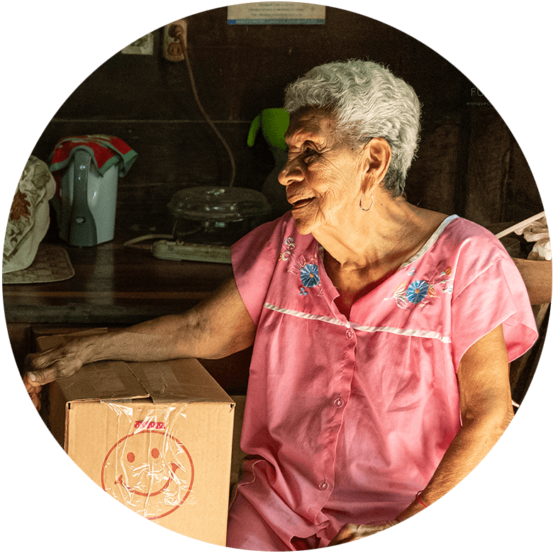 Elderly woman in her rural home with a box of donated supplies, representing how many elders in Cabo Velas face isolation and rely on community support for basic needs.