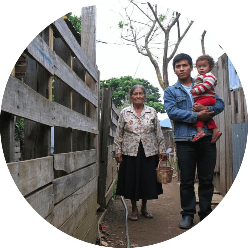 An older woman and a man holding a young child standing in a narrow dirt pathway between wooden fences, representing families in rural communities facing limited resources and support.