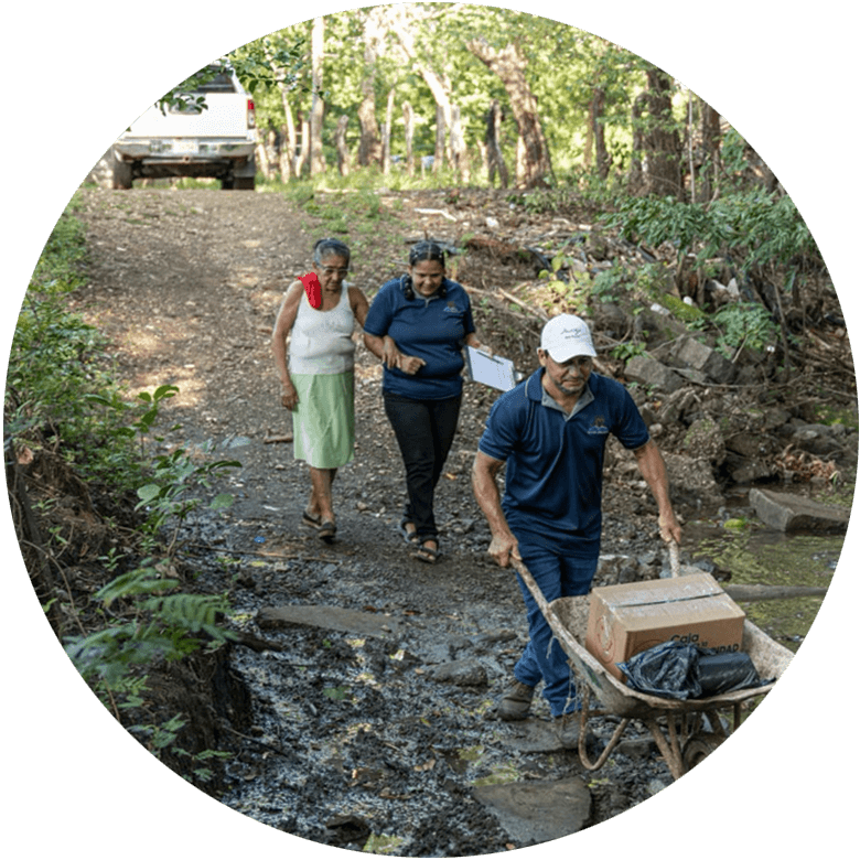 Futuro Brillante team members walking with an elderly woman along a muddy rural path while delivering a box of essential supplies in a wheelbarrow.